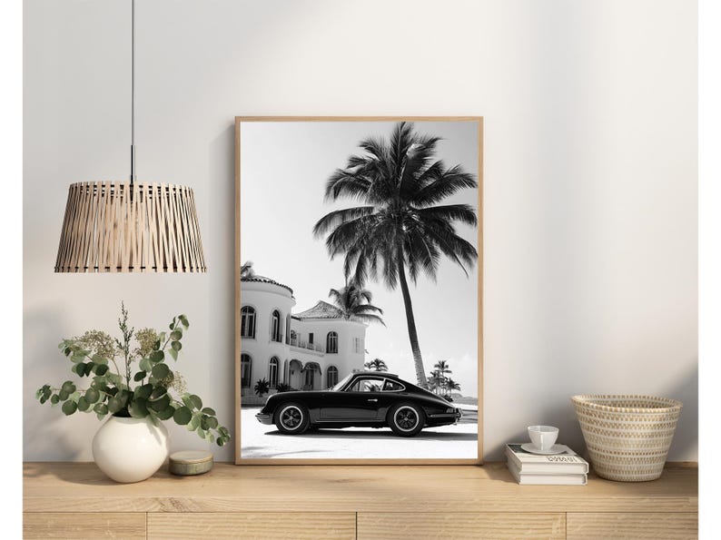 May include: Black and white photograph of a classic car parked in front of a white house with palm trees. The car is a vintage Porsche 911.