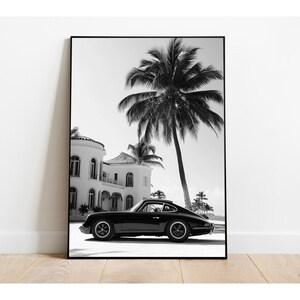 May include: Black and white photograph of a black classic car parked in front of a white house with palm trees in the background.
