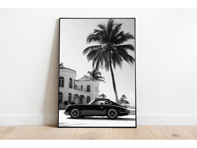 May include: Black and white photograph of a black classic car parked in front of a white house with palm trees in the background.