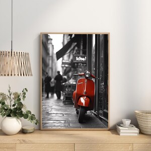 May include: Black and white photograph of a red Vespa scooter parked in a European city street. The scooter is in the foreground and the background is blurred.