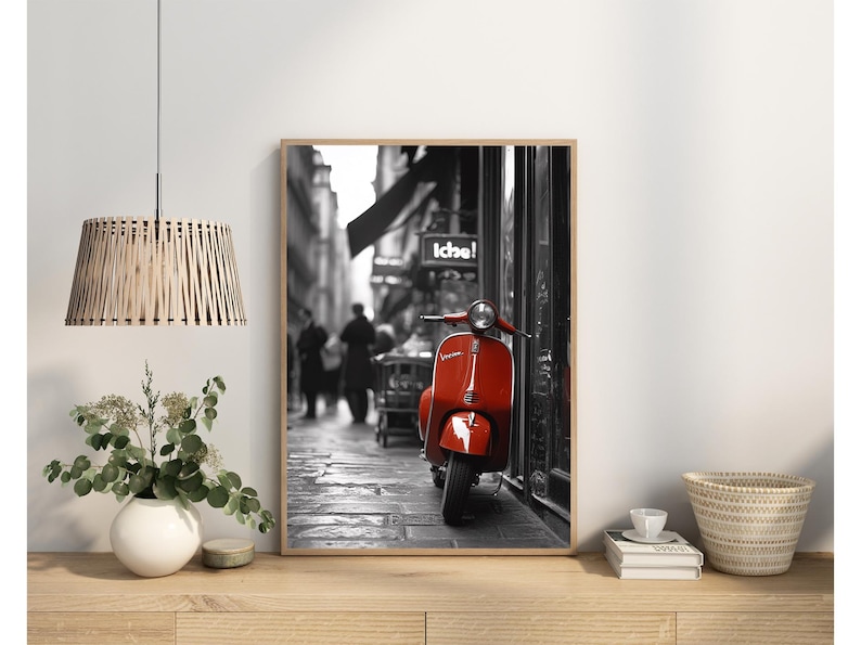 May include: Black and white photograph of a red Vespa scooter parked in a European city street. The scooter is in the foreground and the background is blurred.