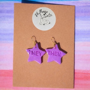 Peut inclure: Deux boucles d'oreilles en forme d'étoile violette avec les mots "THEY THEM" en relief. Les boucles d'oreilles sont suspendues à des crochets dorés. Elles sont présentées sur une carte marron avec un logo circulaire blanc. Le fond est un lavis d'aquarelle bleu, violet et orange.