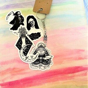 May include: A set of four black and white stickers featuring a skeleton, a woman covering her eyes, a hand reaching for a crescent moon, and a heart-shaped ouija board with the words "Good Bye Hello" inside. The stickers are attached to a silver keychain with a skeleton charm.