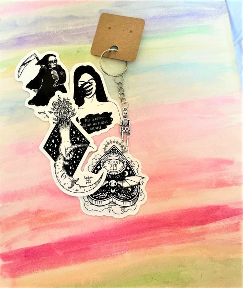 May include: A set of four black and white stickers featuring a skeleton, a woman covering her eyes, a hand reaching for a crescent moon, and a heart-shaped ouija board with the words "Good Bye Hello" inside. The stickers are attached to a silver keychain with a skeleton charm.