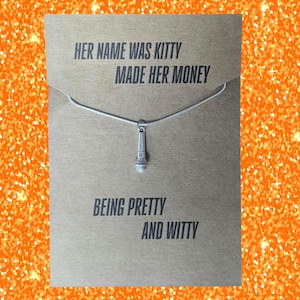 May include: A silver-colored necklace with a microphone pendant hangs on a brown card. The card has the text "HER NAME WAS KITTY MADE HER MONEY BEING PRETTY AND WITTY" in a bold, sans-serif font. The background is a sparkly orange.