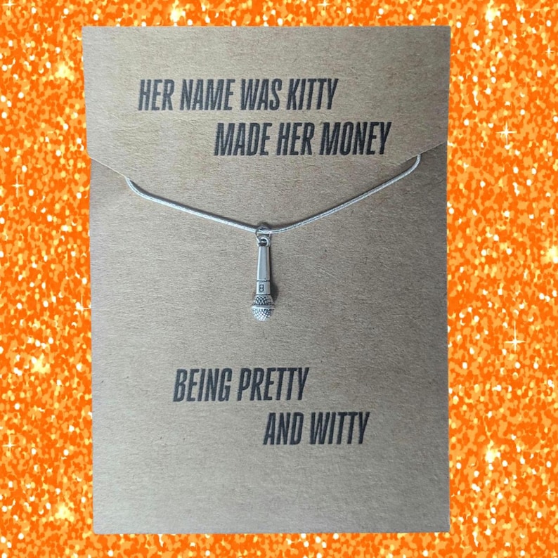 May include: A silver-colored necklace with a microphone pendant hangs on a brown card. The card has the text "HER NAME WAS KITTY MADE HER MONEY BEING PRETTY AND WITTY" in a bold, sans-serif font. The background is a sparkly orange.