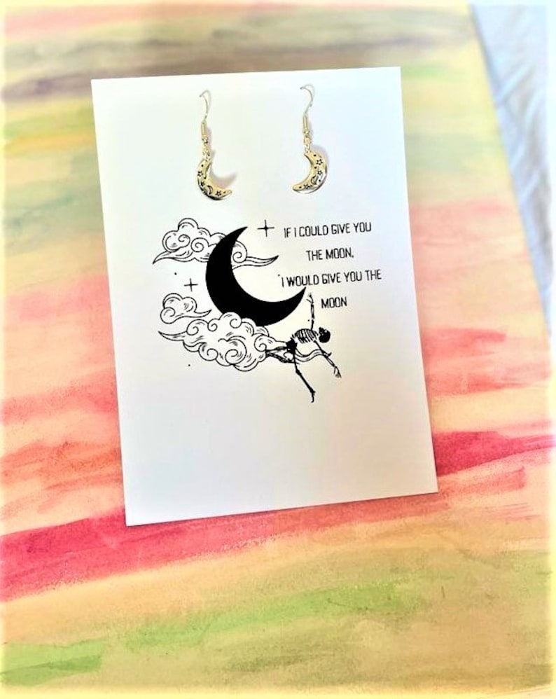 May include: A pair of silver crescent moon earrings with a delicate design. The earrings are displayed on a white card with the text "If I could give you the moon, I would give you the moon."