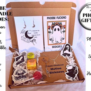 May include: A brown cardboard box filled with a variety of gifts, including a silver moon earring set, two bath bombs, a skeleton keyring, a sticker collection, a silver ghost necklace, a heart-shaped lollipop, and a print of a ghost with the text "Phoebe Fucking Bridgers".
