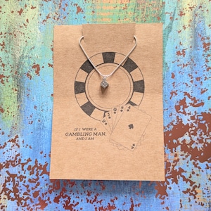 Op de afbeelding: Een zilveren ketting met een kleine, zwarte dobbelsteenbedel. De ketting wordt getoond op een bruine kaart met een pokerchip-ontwerp en de tekst "Als ik een gokker was, en dat ben ik."