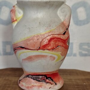 Peut inclure: Un petit vase blanc avec deux anses et une base évasée. Le vase présente un motif marbré avec des tourbillons rouges, jaunes et noirs. Le vase est posé sur une surface en bois.