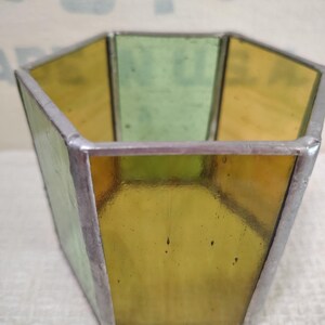 May include: A hexagonal container constructed from panels of translucent yellow and green stained glass joined by silver-toned metal solder lines. The vessel features an open top and flat sides, showcasing a geometric design with visible glass textures.