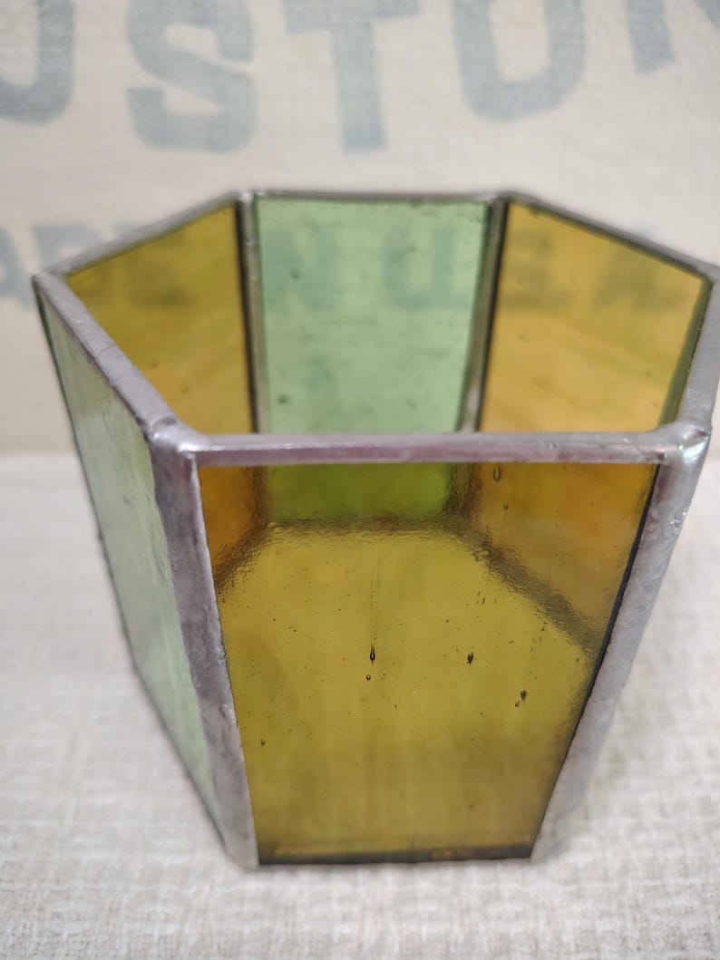 May include: A hexagonal container constructed from panels of translucent yellow and green stained glass joined by silver-toned metal solder lines. The vessel features an open top and flat sides, showcasing a geometric design with visible glass textures.