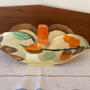 May include: A ceramic serving bowl with a hand-painted design of leaves and fruit in shades of brown, orange, and green. The bowl has a unique shape with two rounded sides and a handle in the center. The handle is a bright orange color.