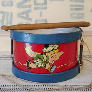 Könnte beinhalten: Eine zylindrische Spielzeugtrommel im Vintage-Stil mit rotem Korpus und blauen Metallrändern. In der Mitte befindet sich eine bunte Illustration eines Kindes in karierter Kleidung, das ein kleines Schwein trägt. Ein Holzschlägel liegt obenauf.