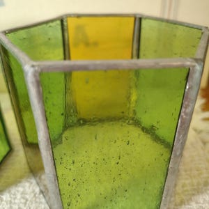 May include: A hexagonal glass container constructed from translucent green and yellow panels joined by silver-toned metal solder lines. The glass features a textured surface with visible air bubbles throughout the material, forming a geometric vessel.