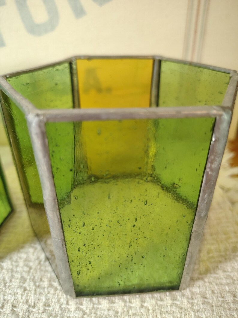 May include: A hexagonal glass container constructed from translucent green and yellow panels joined by silver-toned metal solder lines. The glass features a textured surface with visible air bubbles throughout the material, forming a geometric vessel.