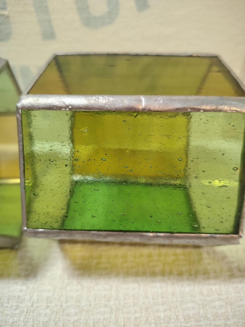 May include: A square-shaped glass box constructed with translucent lime green and yellow panels joined by silver-toned metal solder lines. The glass features visible air bubbles throughout the surface, creating a textured appearance within the frame.