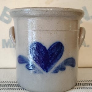 May include: A cylindrical stoneware crock with a light gray finish and two side handles. The front features a hand-painted cobalt blue heart design with decorative leaf-like accents at the base. The rim is thick and rounded with a smooth glazed texture.