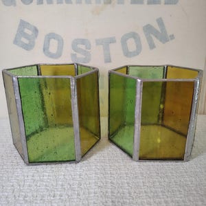 May include: Two hexagonal containers made of green and amber stained glass panels joined by silver-toned metal solder lines. The glass has a textured, slightly translucent appearance. The containers are open at the top and bottom.