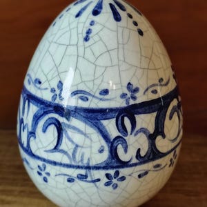 Könnte beinhalten: Ein dekoratives Keramik-Ei mit weißem Grund und blauen Blumen- und Rankenmustern. Das Ei hat einen Craquelé-Effekt und ein blaues Band in der Mitte. Ober- und Unterseite sind mit blauen Akzenten versehen. Ca. 10 cm hoch.