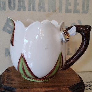 May include: A white ceramic pitcher shaped like a flower with a brown handle and a bee accent. The pitcher has green and red accents on the petals and sits on a small wooden stand.
