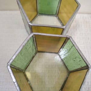May include: Two hexagonal containers constructed from stained glass panels held together by silver-toned metal solder. The panels alternate between translucent yellow and light green glass, creating an open-top geometric vessel design.