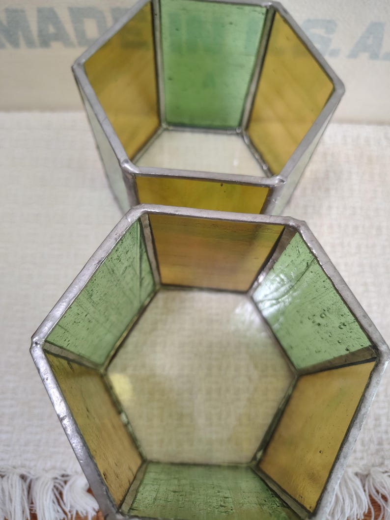 May include: Two hexagonal containers constructed from stained glass panels held together by silver-toned metal solder. The panels alternate between translucent yellow and light green glass, creating an open-top geometric vessel design.