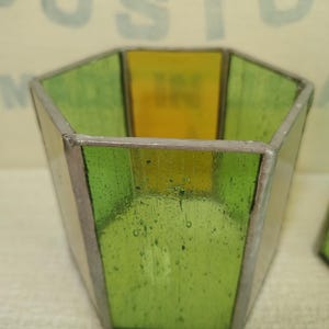 May include: A hexagonal stained glass candle holder featuring alternating panels of translucent lime green and yellow glass. The glass segments are joined together by thin, dark metallic solder lines, creating a geometric, open-topped vessel structure.