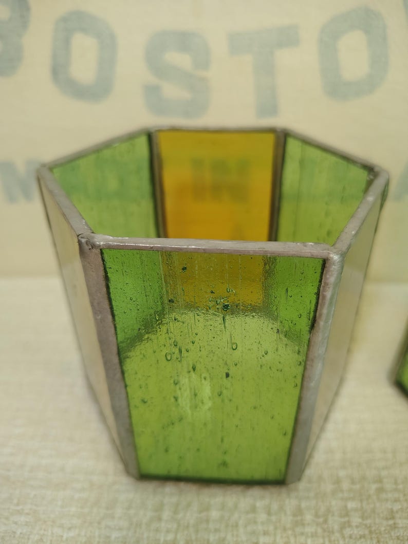 May include: A hexagonal stained glass candle holder featuring alternating panels of translucent lime green and yellow glass. The glass segments are joined together by thin, dark metallic solder lines, creating a geometric, open-topped vessel structure.