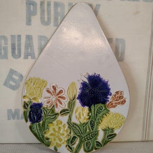 Può includere: Un sottopentola in ceramica a forma di goccia, con sfondo bianco e un colorato motivo floreale. I fiori sono nei toni del giallo, blu, arancione e rosa, con foglie verdi. Utile per proteggere le superfici calde.