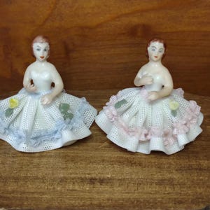 May include: Two porcelain figurines of women in white dresses with detailed lace skirts. One dress has blue trim and the other has pink trim. Each figurine has a painted face and is posed with arms raised.