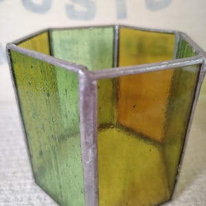 May include: A hexagonal stained glass vessel featuring alternating panels of translucent green and amber glass. The individual glass panes are joined together by thin, dark metal solder lines, creating a geometric container with an open top.