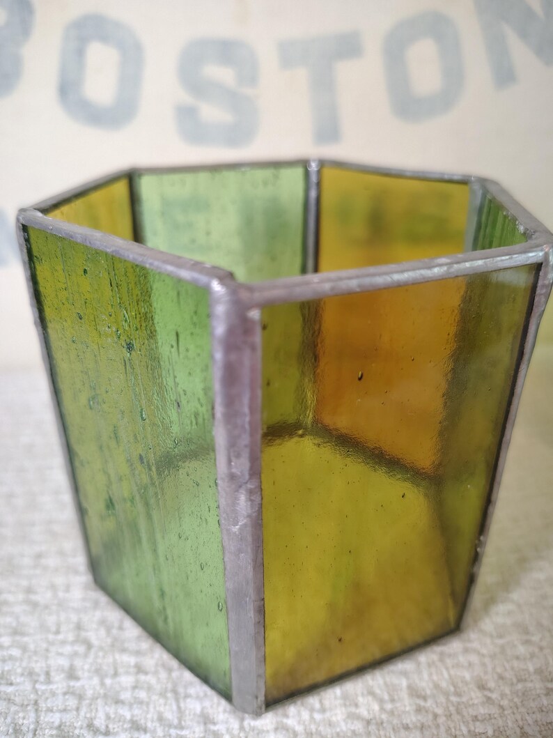 May include: A hexagonal stained glass vessel featuring alternating panels of translucent green and amber glass. The individual glass panes are joined together by thin, dark metal solder lines, creating a geometric container with an open top.