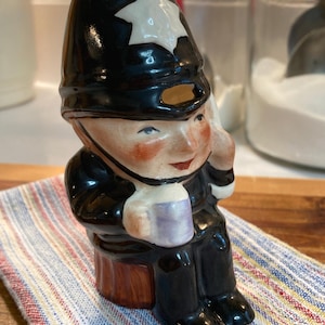 May include: A ceramic figurine of a police officer wearing a black hat with a white star, sitting on a wooden stool and holding a mug. The figurine is painted with realistic details, including a red face and a black uniform.