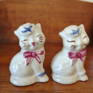 Puede incluir: Dos figuras de gato de cerámica blanca con lazos rosas, detalles azules y ojos cerrados. Cada gato tiene un lazo azul en la cabeza y una pajarita rosa. Se encuentran sobre una superficie de madera.