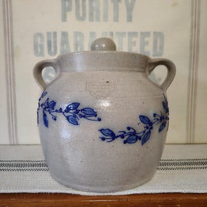 May include: A light gray stoneware crock with two side handles and a matching lid. The exterior features a hand-painted blue vine pattern with leaves and berries wrapping around the center. A stamped maker's mark is visible on the front.