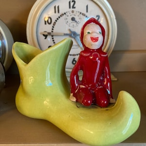May include: A vintage ceramic figurine of a seated figure in a red hooded outfit, atop a light green shoe-shaped planter. A white clock with black numbers is in the background.