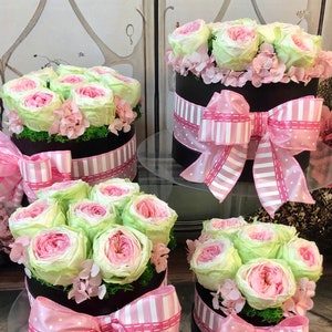 May include: Four black hat boxes with arrangements of pink and green roses. Each box is decorated with a pink and white striped ribbon and a large bow. The flowers are surrounded by green moss.