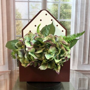 May include: A brown envelope-shaped planter with a white polka dot pattern on the front. The planter is filled with green hydrangea flowers and ferns.