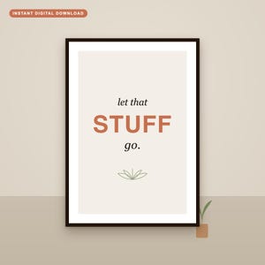 May include: A framed wall art print featuring a cream-colored background with the black text let that and the orange text STUFF followed by the black text go. Below the text is a simple green line drawing of a lotus flower or plant icon.