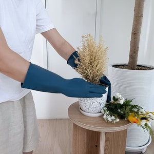 May include: A person wearing blue rubber gloves is repotting a small plant with white speckled pot. The plant is in a white pot with a white speckled design. The person is standing next to a large white pot with a plant inside.