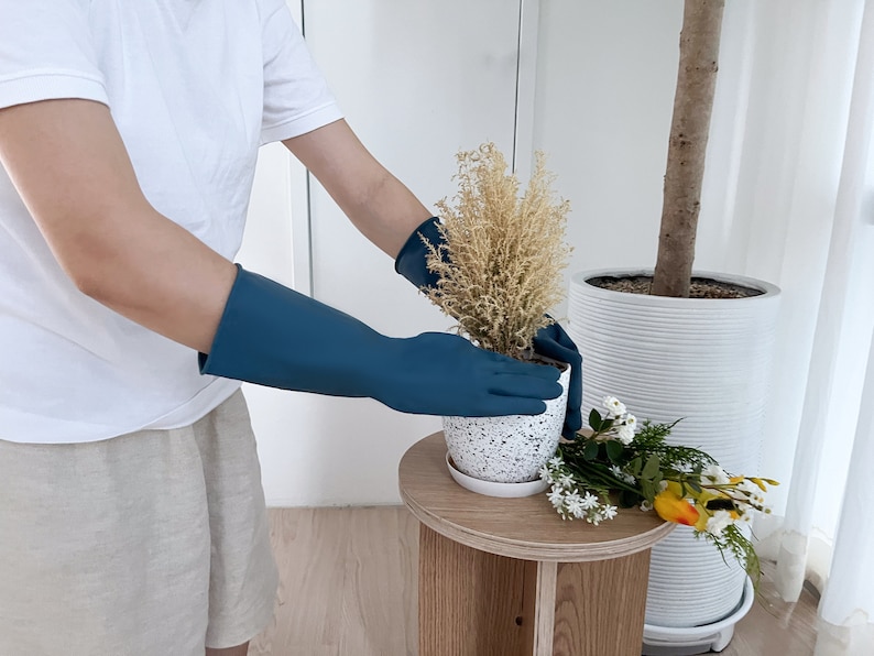 May include: A person wearing blue rubber gloves is repotting a small plant with white speckled pot. The plant is in a white pot with a white speckled design. The person is standing next to a large white pot with a plant inside.