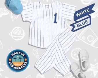 May include: White and blue striped baseball uniform with a white jersey and blue pants. The jersey has a number "1" on the front. The uniform is laid out on a gray background with a blue baseball cap, white sneakers, and a patch that says "Made in Los Angeles, California".