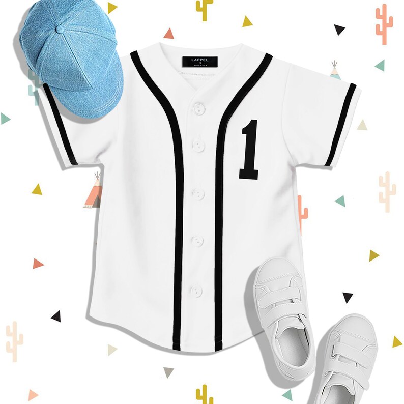 Op de afbeelding: Een wit baseballshirt met zwarte bies en het nummer "1" op de voorkant. Het shirt ligt plat op een witte achtergrond met een blauwe baseballpet en witte sneakers.