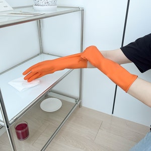 May include: A person wearing orange rubber gloves is cleaning a metal shelf with a white cloth. The shelf has a glass top and a glass bottom shelf. There is a glass of red liquid on the bottom shelf.