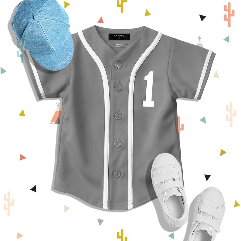 Op de afbeelding: Een grijs baseballshirt met witte strepen en een wit nummer '1' op de voorkant. Het shirt heeft een knoopsluiting aan de voorkant en korte mouwen. Een blauwe denim baseballpet en witte sneakers zijn ook afgebeeld.