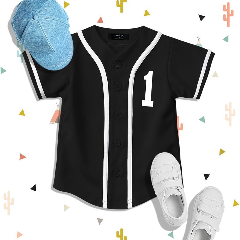 Op de afbeelding: Een zwart baseballshirt met witte bies en het nummer "1" op de voorkant. Het shirt wordt gecombineerd met een lichtblauwe baseballpet en witte sneakers.