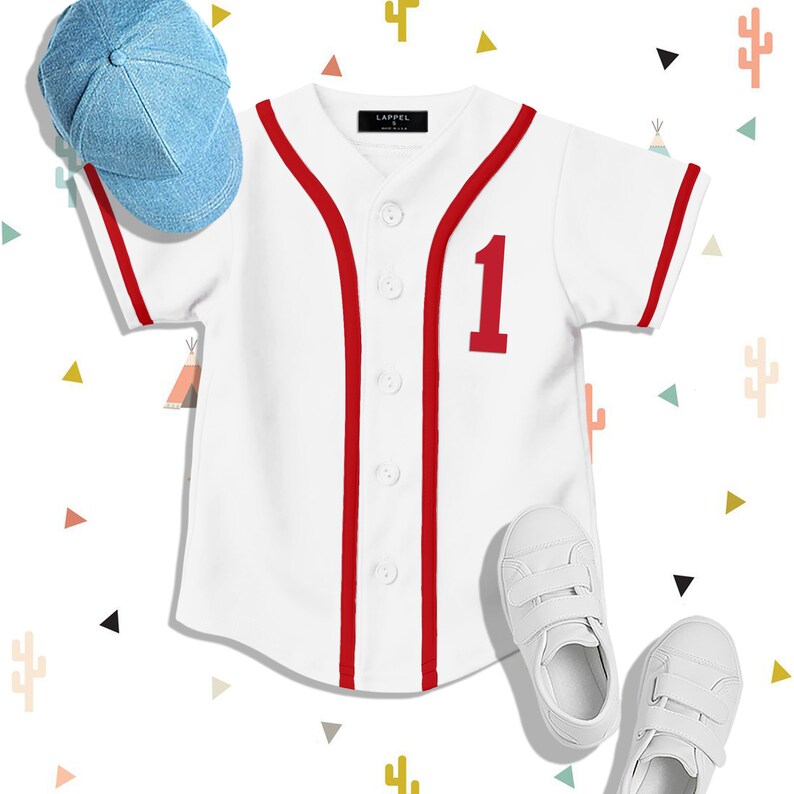 Op de afbeelding: Wit baseballshirt met rode bies en het nummer "1" op de voorkant. Het shirt ligt plat op een witte achtergrond met een blauwe baseballpet en witte sneakers.
