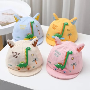 Puede incluir: Cuatro gorras de béisbol para niños con diseños de dinosaurios. Cada gorra tiene un color diferente: azul, amarillo, beige y rosa. Las gorras tienen el lema 'Never Stop' y orejas de dinosaurio.