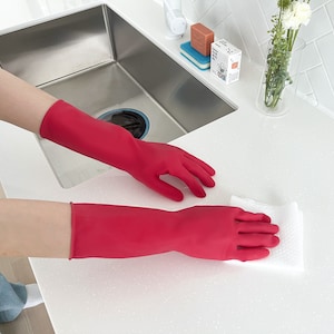 May include: A person wearing red rubber gloves is cleaning a white countertop with a white cloth. The countertop is next to a stainless steel kitchen sink.